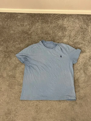 Blå t-shirt från Polo Ralph Lauren - Snygg ljusblå t-shirt från Polo Ralph Lauren med klassisk rund hals och korta ärmar. T-shirten har en liten mörkblå Polo-logga broderad på bröstet. Tillverkad i mjuk bomull som är skön mot huden. Perfekt för en chill och stilren look. Tröjan är använd så slitage finns 