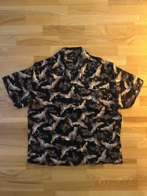 Svart Amiri skjorta med fågelmönster - Säljer en svart skjorta från Amiri i storlek small med korta ärmar och knappar framtill. Skjortan har ett detaljerat mönster med vita och beige fåglar och grenar, vilket ger en unik och cool vibe. Materialet är mjukt och lyxigt, perfekt för dig som gillar statement-plagg.