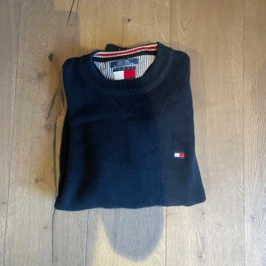 Mörkblå stickad tröja Tommy Hilfiger - Snygg mörkblå stickad tröja från Tommy Hilfiger i storlek L. Tröjan har rund hals med ribbad kant och en liten broderad logga på bröstet. Materialet är mjuk bomull och insidan av halsen har randiga detaljer i rött, vitt och blått.