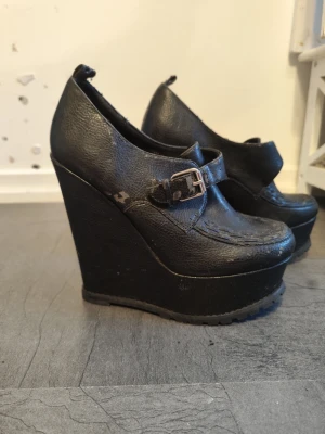 Svarta wedge skor från Tiamo, strl 40 - Säljer ett par svarta wedge skor från Tiamo i storlek 40. Skorna har en hög kilklack, rund tå och ett brett spänne över foten. Materialet är skinnimitation med synliga sömmar som detalj. Perfekt för dig som gillar att sticka ut med höga klackar.