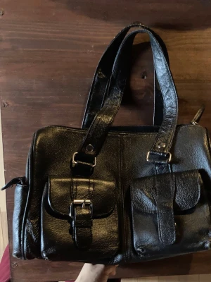 Svart axelväska - Mått: 50 x 23 cm. Är lite sliten på hörnen (syns på bild 7), har lite rosa färg på ena sidan (bild 4) och är lite nött på framsidan där handtagen sitter fast. Rekommenderar att inte ha allt för tunga föremål i väskan, utöver detta är den mycket fin och har många små fickor som antingen öppnas med knapp, dragkedja eller magnet knapp 