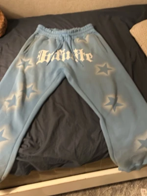 Blå mjukisbyxor med stjärnor och tryck - Snygga ljusblå mjukisbyxor från Bandite med vita stjärnor och stort tryck framtill. Byxorna har snörning i midjan och är tillverkade i mjukt bomullsmaterial. Perfekta för chill och streetwear. Fickor på sidorna och bak för extra funktion. Säljer för behöver pengar