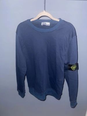 Blå långärmad tröja från Stone Island - Säljer en snygg marinblå långärmad tröja från Stone Island med den klassiska logotypen på vänster ärm. Tröjan har rund hals och ribbade muddar vid ärmslut och nederkant. Perfekt för dig som gillar streetwear och vill ha något stilrent.