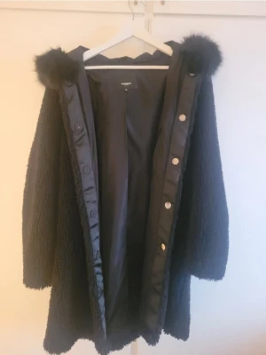 Svart joplin jacka med päls från Rockandblue - Svart joplin jacka från Rockandblue med fluffig huva och pälsdetaljer. Jackan har långa ärmar, knappar framtill och är tillverkad i ett mjukt syntetmaterial. Perfekt för kalla dagar när du vill ha en cool och mysig look.