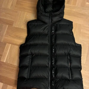 Svart dunväst från Moose Knuckles - Säljer en svart dunväst från Moose Knuckles i storlek M. Västen har huva, två dragkedjeförsedda fickor och en quiltad design. Perfekt för kyliga dagar och har en stilren look med logga längs dragkedjan framtill. Köpt på NK Göteborg. Hör av dig om du har frågor!