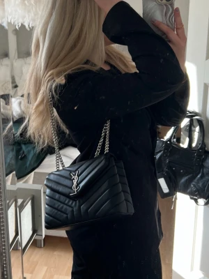 Svart quiltad handväska från YSL - Säljer en svart quiltad handväska från YSL med silverfärgad kedja och ikonisk YSL-logga framtill. Väskan har en stilren, strukturerad form och är tillverkad i skinn. Perfekt för dig som vill ha en lyxig accessoar med snygga detaljer. Har fått den 
