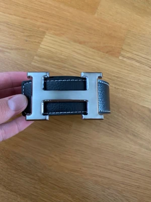 Svart skinnbälte med H-spänne - Snyggt svart bälte i skinn med ett stort, silvrigt H-format metallspänne. Bältet har vit kontrastsöm längs kanterna och en stilren, klassisk look. Perfekt för att ge outfiten en extra touch av lyx och attityd.