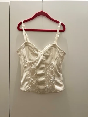 Champagne/vit färgad korsett topp med spets - Säljer en supersnygg champagne/vit färgad korsett med justerbara axelband. Toppen har snygga genomskinliga spetsdetaljer framtill och är tillverkad i glansigt satinliknande material. Kuporna är formade och ger en feminin siluett. Perfekt som en festtopp att klä upp eller ned!🥰🥰Från H&M men lappen bortklippt. I mycket bra skick men med några nopprar här o där som knappast syns.