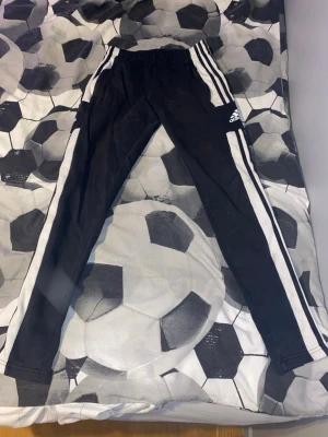 Svarta Adidas mjukisbyxor XS - Svarta mjukisbyxor från Adidas i storlek XS med vita ränder längs sidorna och klassisk Adidas-logga på benet. Byxorna har elastisk midja och är gjorda i mjukt material, perfekta för chill eller sport. Primegreen-kvalitet.