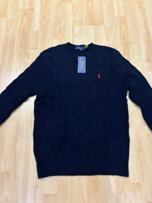 Mörkblå stickad tröja Polo Ralph Lauren - Snygg mörkblå stickad tröja från Polo Ralph Lauren med klassiskt kabelmönster och rund hals. Tröjan har lång ärm och den ikoniska röda Polo-loggan broderad på bröstet. Perfekt för dig som gillar stilrena och tidlösa plagg. Bra passform 