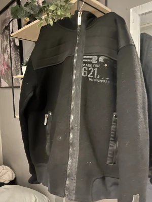 Svart zip hoodie - Tröjan är en svart zip-hoodie. Den har ett stort silvergrått tryck på bröstet med texten “RG 512” och ett mönster som liknar vingar bakom texten. Under loggan står orden “Being part of it.  Den har dragkedja hela vägen ner och två sidofickor som också stängs med dragkedja. Tyget ser mjukt och lite stretchigt ut, som klassiskt hoodie-material.