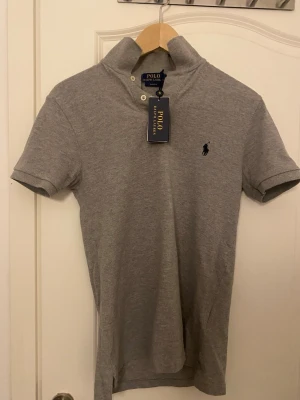 Grå polotopp från Polo Ralph Lauren oanvänd - Säljer en stilren grå polotopp från Polo Ralph Lauren i slim fit. Toppen har korta ärmar, klassisk krage med två knappar och den ikoniska broderade loggan på bröstet. Materialet är mjuk bomull som känns skönt mot huden. Perfekt för dig som gillar enkel och snygg design.