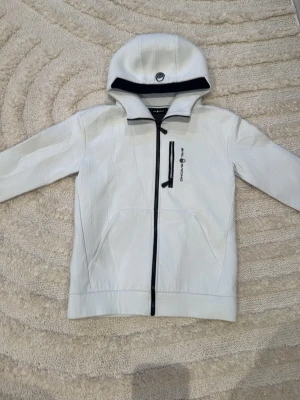 Vit vindjacka från Sail Racing - Säljer en stilren vit vindjacka/ kofta från Sail Racing med svart dragkedja och logga på bröstet. Jackan/ koftan har huva, en bröstficka med dragkedja och två sidofickor. Materialet är lätt och känns sportigt, perfekt för alla dagar. Passformen är normal och jackan har långa ärmar.