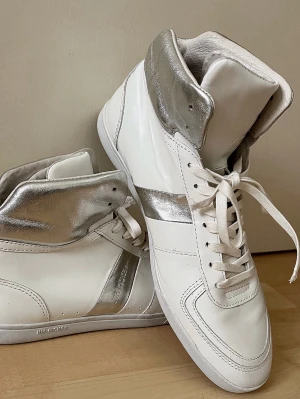 Vita och silver high top sneakers Jim Rickey - Snygga high top sneakers från Jim Rickey i vitt skinn med metallic-silver detaljer runt ankeln och på sidan. Klassisk snörning framtill och rund tå. Perfekta för dig som vill sticka ut med en clean men ändå unik look. Sparsamt använda, endast tecken på användning syns på sulan. Finns ej att få tag på idag. 