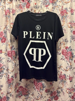 Svart t-shirt från Philipp Plein - Svart t-shirt från Philipp Plein med stor vit logotyp och tryck framtill. Klassisk rund hals och korta ärmar. Materialet är mjuk bomull med lite stretch från lycra, vilket gör den skön och följsam. Perfekt för dig som gillar statement-plagg och streetstyle.