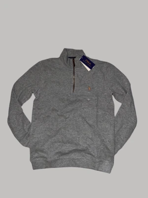 Grå half zip tröja från Polo Ralph Lauren - Snygg grå långärmad tröja från Polo Ralph Lauren med half zip och klassisk krage. Tröjan har ett broderat logomärke på bröstet och är tillverkad i mjuk bomull. Perfekt för dig som gillar stilrena och bekväma plagg med premiumkänsla.
