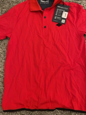 Röd Replay Evoflex piké t-shirt - Snygg röd piké t-shirt från Replay Evoflex med klassisk krage och knappar framtill. T-shirten har ett svart Replay-märke på bröstet och är gjord i ett stretchigt syntetmaterial som känns sportigt och modernt.