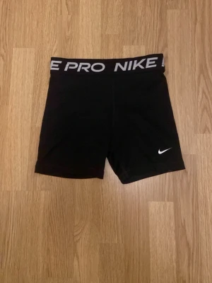 Svarta Nike Pro träningsshorts - Nike Pro shorts i svart med vit logga och bred resår med Nike Pro-tryck. Tight passform och tillverkade i stretchigt syntetmaterial som andas, perfekta för gym eller sport. Klassisk design med korta ben och hög midja.