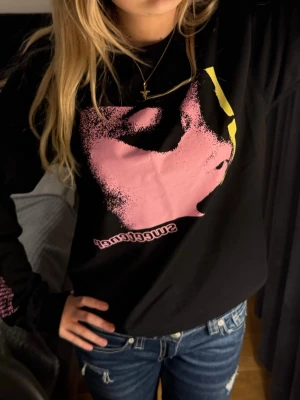 Ariana Grande Sweetener långärmad tröja - Säljer en svart långärmad tröja från Ariana Grande Sweetener tour från H&M med stort rosa tryck av ett ansikte på framsidan, gul text 'thank u, next' på ena ärmen och rosa turnéstäder på andra ärmen. Tröjan har en cool, oversized vibe.
