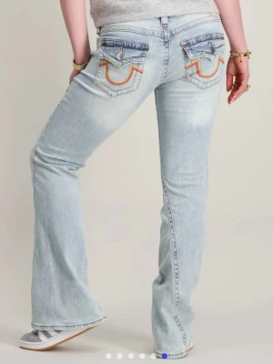 True Religion ljusblå bootcut jeans - Säljer ett par ljusblå jeans från True Religion med bootcut passform och låg midja. Jeansen har snygga fickdetaljer med broderade sömmar i orange och rött samt silverfärgade knappar på bakfickorna. De är jättefina men är tyvärr för stora så de ska egentligen sitta tight. De har lite svart färg på framsidan som går bort i tvätten. Några lösa trådar här och där men de syns absolut inte och går lätt att klippa bort. Knappt använd annars och ser nya ut. Säg till om du har några frågor💕