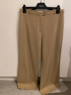 Kostymbyxor  - Snygga beiga kostymbyxor från Aleyna Collection i storlek 48. Byxorna har en wide passform med raka ben, hög midja och klassisk knapp- och dragkedjestängning. Materialet är 67% polyester och 33% viskos, vilket ger en mjuk och följsam känsla. Använda en gång innan.