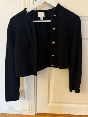 Svart stickad kofta från H&M - Säljer en svart stickad kofta från H&M i storlek S. Koftan har ribbad struktur, guldfärgade knappar framtill och två dekorativa fickor. Modellen är lite croppad och har långa ärmar.