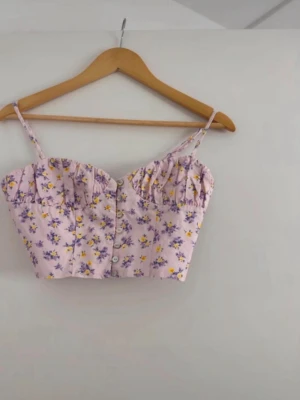 Blommig croppad linnetopp från Zara - Superfin croppad linnetopp från Zara i ljuslila med blommigt mönster i gult och lila. Toppen har smala axelband, knappar framtill och smockad rygg för extra komfort. Perfekt för varma dagar och sommarhäng! Oanvänd 