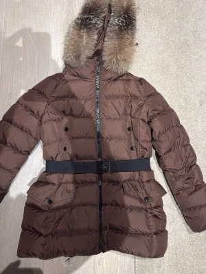 Brun Moncler dunjacka med päls storlek 3 - Säljer en brun Moncler dunjacka med fluffig päls runt huvan och snyggt bälte i midjan. Jackan har dragkedja, knappar och två stora fickor framtill. Perfekt för kalla dagar med sin quiltade design och långa ärmar. Storlek 3 