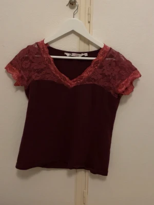 Vinröd topp med spets från Instinct - Supersnygg vinröd topp från Instinct i storlek M. Toppen har korta ärmar och v-ringning med dekorativ spets i en matchande röd nyans över axlar och halsringning. Perfekt för dig som vill ha något lite extra till outfiten.🙏🥰