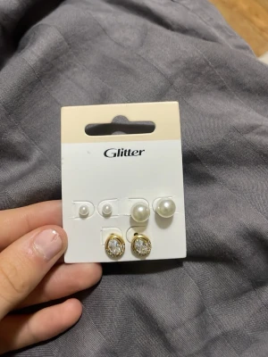 Ingen väst på bilderna - Bilderna visar ett set med örhängen från Glitter, inklusive pärlörhängen och guldpläterade örhängen med glittrande stenar. Ingen väst finns med på bilderna.