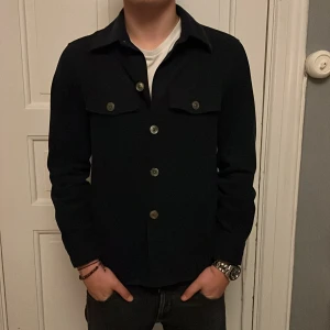  overshirt från Oscar Jacobson - Stilren marinblå overshirt från Oscar Jacobson med klassisk krage, raka linjer och fyra knappar framtill. Två bröstfickor med lock och knappar ger en snygg touch. Perfekt att bära över en t-shirt för en clean look. Nypris 3000kr