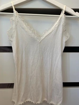 Vit ribbad linnetopp med spets från Amisu - Säljer en vit ribbad linnetopp från Amisu i storlek XS. Toppen har tunna axelband och v-ringning med dekorativ spets längs kanterna. Materialet är mjukt och lätt, perfekt för varma dagar. Snyggt vågig nederkant för extra detalj.