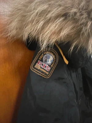 Svart Parajumpers parkajacka med päls - Svart parkajacka från Parajumpers med stor huva och fluffig pälsdetalj. Jackan har flera fickor, dragkedja och coola patchar på ärmen. Insidan har etikett med 'Masterpiece Series'. Perfekt för kalla dagar och riktigt snygg streetstil.