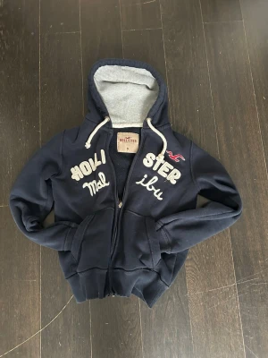 Hollister Hoodie - En snygg marinblå hoodie från märket Hollister med snygga detaljer på framsidan. Skriv om du har frågor! Pris kan diskuteras.