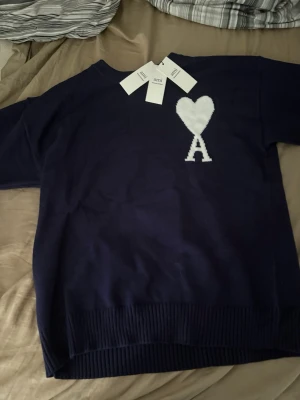Mörkblå stickad tröja från Ami - Säljer en mörkblå stickad tröja från Ami i storlek S. Tröjan har ett vitt hjärta med bokstaven A på bröstet och ribbade muddar vid ärmslut och nederkant. Materialet är mjuk bomull och passformen är loose. Perfekt för dig som gillar stilrena detaljer.