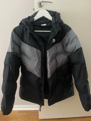 Svart och grå dunjacka från Nike - Nike pufferjacka i svart och grått. Jackan har huva, dragkedja framtill och är tillverkad i syntetmaterial. 