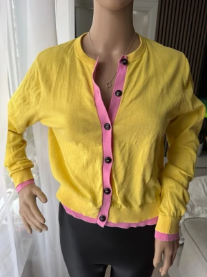 Gul och rosa kofta med knappar ZARA  - Snygg gul kofta med rosa detaljer längs knappraden, nederkant och ärmslut. Tröjan har svarta knappar framtill och är långärmad. Perfekt för dig som vill ha en färgklick i garderoben. Tillverkad i mjuk bomull som känns skön mot huden.