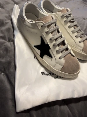 Golden Goose Superstar sneakers - Golden Goose Superstar sneakers i vitt skinn med grå mockadetaljer på tungan och grå snörning. Sidan har en svart stjärna i mocka och GGDB/SSTAR-tryck i guld. Platt sula med svart mönstrad undersida och silverfärgad häl. Snygg och unik design för dig som gillar streetstyle.