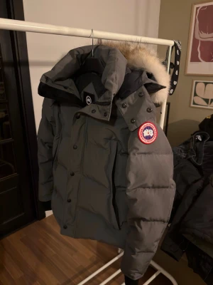 Grå Canada Goose Wyndham dunjacka med päls - Säljer en grå dunjacka från Canada Goose med stor fluffig päls på huvan. Jackan har svarta detaljer, dragkedja och knappar framtill samt märkets ikoniska patch på ärmen. Perfekt för kalla vinterdagar och riktigt snygg stil. Användes knappt 1 vinter så i princip ny! Bästa batchen!