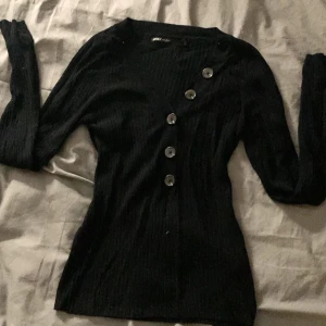 Svart ribbad kofta från ONLY - Snygg svart ribbad kofta från ONLY med v-ringning och fem stora knappar framtill. Koftan har lång ärm och är figurnära i modellen, perfekt att styla med jeans eller kjol. Materialet är mjukt och stretchigt, vilket gör den bekväm att bära hela dagen.