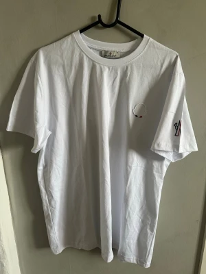 Vit Moncler t-shirt med broderi - Vit t-shirt från Moncler i mjuk bomull med rund hals och korta ärmar. På bröstet finns ett broderat Moncler-emblem i vitt, rött och svart, och på ena ärmen syns en broderad Moncler-logga. Perfekt för dig som gillar stilrena designer och exklusiva märken.