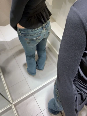 True Religion blå bootcut jeans - Säljer ett par blå jeans från True Religion med klassiska kontrastsömmar och broderade bakfickor. Modellen är lågmidjad med bootcut och har slitningar framtill. Jeansen är i denim med detaljerade fickor och logopatch bak.