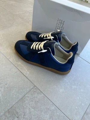 Maison Margiela sneakers blå - Maison Margiela sneakers i marinblått med vita snören och beige insida. Skorna har en kombination av mocka och skinn, samt en klassisk gummisula. Snygg retrostil med rund tå och platt sula, perfekt för dig som gillar stilrena detaljer.