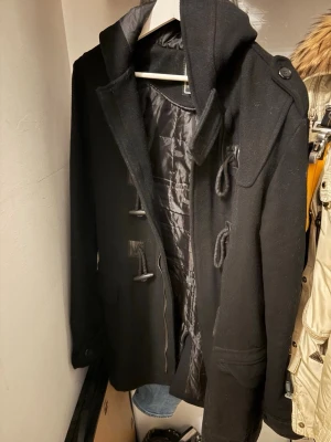 Svart rock med snörning och huva - Svart rock med huva och snygga snörningsdetaljer framtill. Jackan har en klassisk form med stora fickor och fodrad insida för extra värme. Perfekt för dig som vill ha en stilren och enkel look under kyliga dagar.