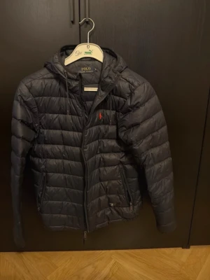 Svart pufferjacka Polo Ralph Lauren - Säljer en svart pufferjacka från Polo Ralph Lauren i storlek S. Jackan har huva, dragkedja framtill och två sidofickor med dragkedja. Klassisk quiltad design och den ikoniska röda loggan på bröstet. Perfekt för kyliga dagar och riktigt snygg till vardags.