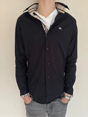 Svart Burberry skjorta med rutig detalj - Riktig fet burberry skjorta i svart. Stl S och sitter bra. Modellen är 184cm. Skick |8/10| och har inga tydliga defekter. Pris 699kr och nypris ca4900kr så passa på!🤩