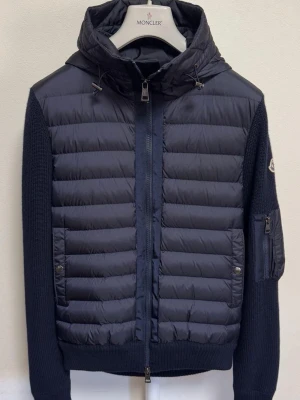 Moncler Cardigan SITTER SOM MEDIUM - Snygg mörkblå pufferjacka från Moncler i storlek XL. Jackan har stickade ärmar, huva med snörning och dragkedja framtill. Fickor med tryckknappar och en extra ficka med dragkedja på ärmen. Perfekt för dig som vill ha en cool och varm jacka. Passar cm omkring 180cm