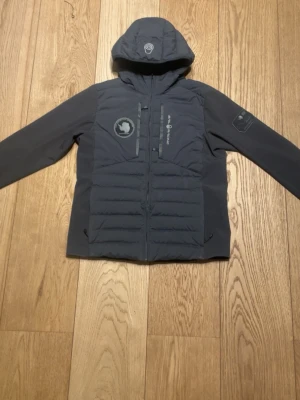 Mörkgrå jacka från Sail Racing - Säljer en mörkgrå pufferjacka från Sail Racing med huva och snygga detaljer som patch på bröstet och logga på ärmen. Jackan har dragkedja framtill och quiltad framsida. Perfekt för dig som gillar stilren och sportig look.