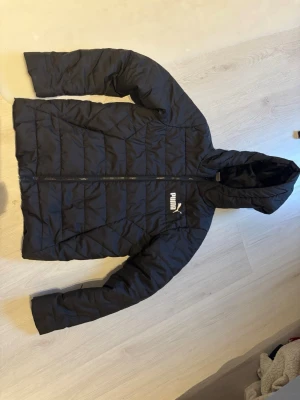 Svart pufferjacka från Puma - Svart pufferjacka från Puma med huva och diskret logga på bröstet. Jackan har quiltad design, dragkedja framtill och är tillverkad i syntetmaterial. Perfekt för kalla dagar när du vill hålla dig varm och samtidigt se sportig ut. Passar från stl 146