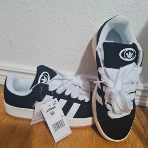 Adidas Campus sneakers svart/vit - Adidas Campus sneakers i svart mocka med klassiska vita ränder och tjocka vita snören. Låg modell med rund tå och platt sula. Ikonisk Adidas-logga på plösen och insidan. Perfekt för dig som gillar retrostil och streetwear.